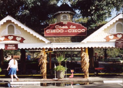 Photo of a multi-gabled roof shading a waiting area, with a sign proclaiming CASA DE CHOO-CHOO. Down on the sidewalk, a pink cartoon cat sits up proudly.