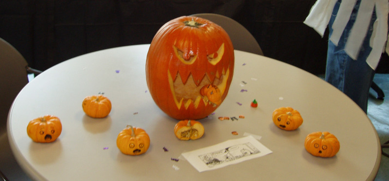A regular-sized jack-o-lantern with a gaping, fanged mouth. Around it on the table are a whole bunch of tiny pumpkin-like squash with scared faces drawn on them in marker. One has been split in two, with one piece stuck in the large one's mouth. A piece of paper with a comic strip is sitting on the table, too small to make out any of the images or words.