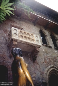 Looking up past a bronze statue of a woman, seen from behind, toward an ornately carved balcony set in the wall of a stone building with three levels or peaked windows and doors. Vines trail from a wooden walkway the next level up from the balcony.