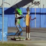 A pair of sculptures made of metal car parts like exhaust pipes and engine parts. One is a very cylindrical man with a short beard and mustache and a finned hat like a conquistador-era Spanish soldier and holds a spear. Bienvenidos is written on a shield. The other is crouching slightly as if to jump, holding its arms out, and has an elongated head painted green with a serrated ridge along the top, curved hoses sprouting from the sides, and a fish-like face with bulbous red eyes. A cinder block wall next to them is painted blue, as is a corrugated metal building behind them.