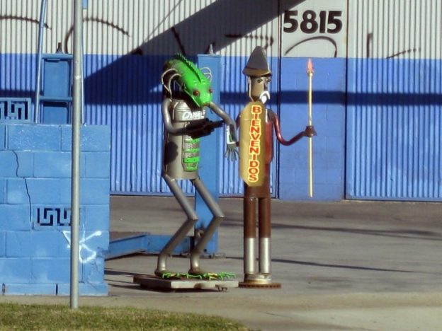A pair of sculptures made of metal car parts like exhaust pipes and engine parts. One is a very cylindrical man with a short beard and mustache and a finned hat like a conquistador-era Spanish soldier and holds a spear. Bienvenidos is written on a shield. The other is crouching slightly as if to jump, holding its arms out, and has an elongated head painted green with a serrated ridge along the top, curved hoses sprouting from the sides, and a fish-like face with bulbous red eyes. A cinder block wall next to them is painted blue, as is a corrugated metal building behind them.