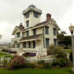 It's a cloudy day. Behind a white picket fence and green lawn, there's a whitewashed, gabled housel Most of it is two stories, but a square tower in the middle rises two more levels, and the tower is topped with a railing above which which you can just glimpse the top of a glass structure.