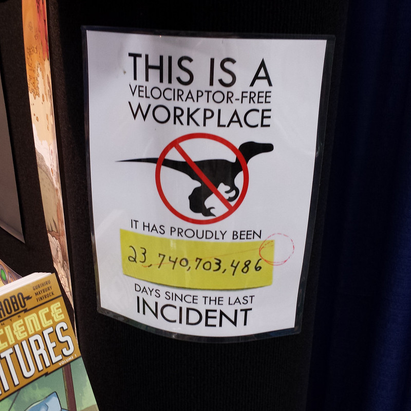 Paper sign on a wall: THIS IS A VELOCIRAPTOR-FREE WORKPLACE. It has proudly been ____ days since the last incident. (23,740,703,486 has been written in by hand, then crossed out with a 0 written in.)