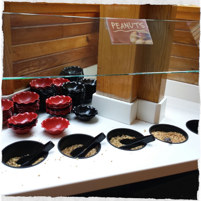 A shelf with stacks of ice cream bowls and bins with toppings including chopped peanuts. There are peanuts scattered all over the shelf.