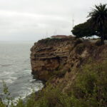 It's gloomy. The sky and sea are almost the same shade of gray. Looking along a steep cliffside toward a rock outcropping. It's high, maybe ten times as high as the low building perched on top of it, and grooves show dozens of layers stacked from the sea below to a bit below the top.