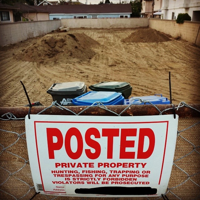 Sign in front of a house-sized empty lot with only dirt, surrounded by walls and other houses. Posted: No hunting, fishing or trapping...