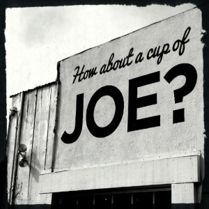 Stark black and white image of a sign painted directly on an outer wall. Wooden boards are visible next to it, and the top of a window frame below it. The sign reads "How about a cup of" in cursive, and then in MUCH LARGER SANS-SERIF BLOCK LETTERS, "JOE."