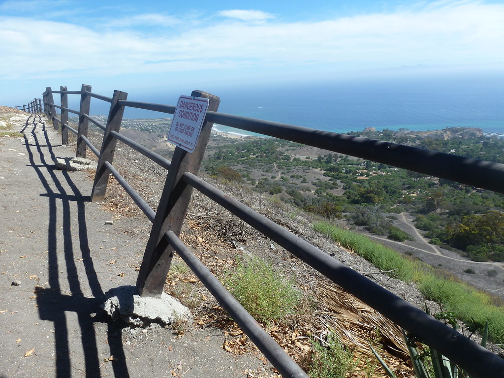 Post-and-rail fence at the top of a cliff. The ground has eroded around the concrete holding the posts in place, and the fence is leaning toward the empty space. A signs warns DANGEROUS CONDITION.