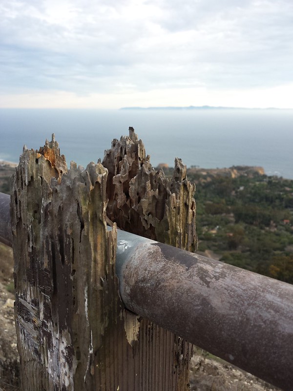 Horribly-termite-eaten wooden post with a metal bar running through a trough. Land beyond it is clearly a lot lower.