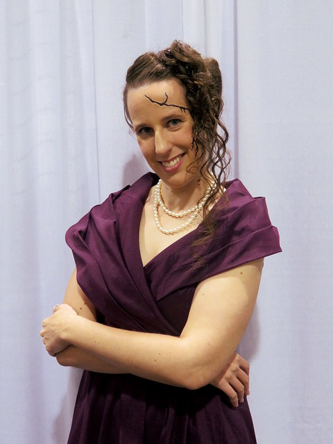 Whitney Frost Cosplay: Woman in a 1940s style purple dress, spidery cracks running down the side of her face, and hair with half a victory roll and curls on one side.