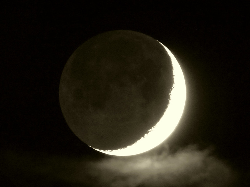 Overexposed crescent moon, with the  night side faintly visible and a glowing cloud below it.