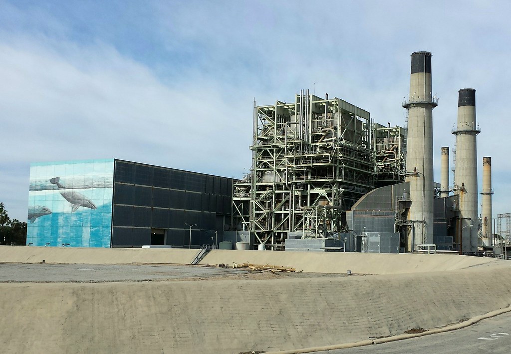 Industrial building with smokestacks and scaffolding. Next to it is a boxy building painted with an ocean scene featuring whales.
