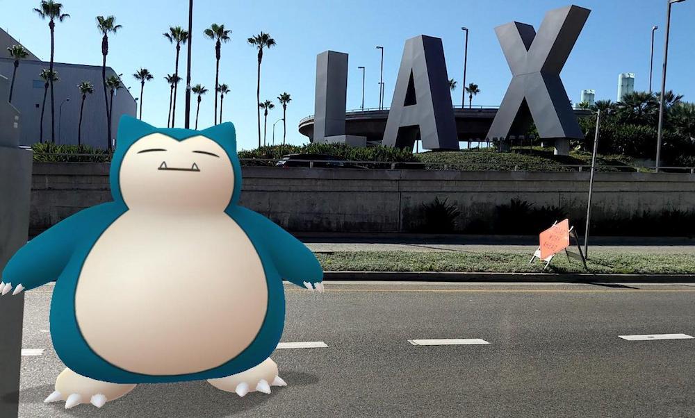 A Snorlax (large fictional creature with a white face and belly, and blue around the edges and on its limbs, with cat ears and eyes closed. It looks sort of like Totoro if he couldn't quite stay awake.) superimposed on the road in front of a giant free-standing LAX sign.
