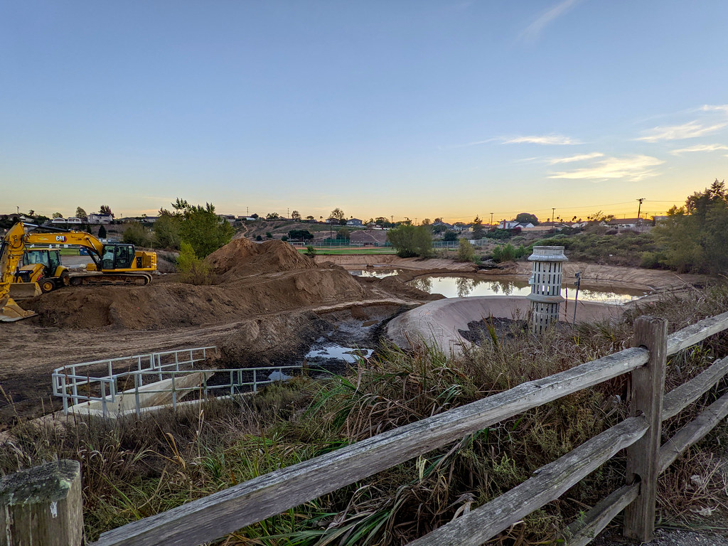 The same lake and drain, but the plants are mostly brown and yellow, the water's a lot lower and smaller, and construction equipment is moving dirt around.