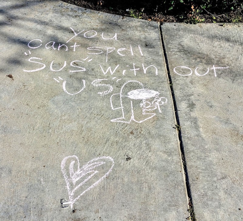 Chalk writing on sidewalk: You can't spell 'sus' without 'us'. Drawings of a heart and an Among Us crewmate holding a flower.