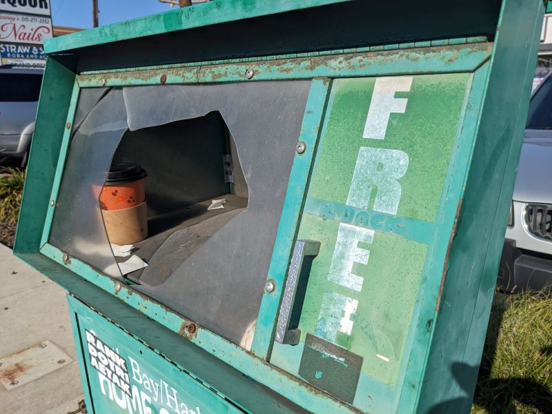 Newspaper vending box labeled FREE. The glass panel on the front is broken, and a cardboard coffee cup sits inside it.