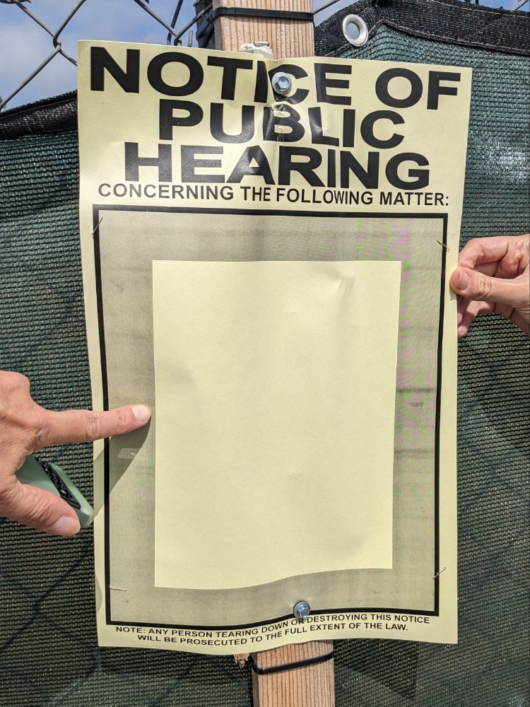 Paper sign on a chain link fence, NOTICE OF PUBLIC HEARING with a big empty space where an 8½×11" page should be, but isn't.