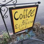 A squareish hand-painted metal sign with the words 'Coffee,' 'Espresso' and 'Wifi' stacked vertically. The letters and border are black on a bright yellow background. The sign hangs from an ornamented wrought-iron-style stand above a planter and is viewed diagonally from above. Between 'Coffee' and 'Espresso' there's a drawing of a cup and saucer with steam rising from it.