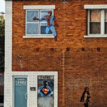 A red-brown brick wall. Two sets of windows are upstairs, and a door downstairs. Next to the door is a painted silhouette of a man pulling his outer shirt open and revealing the Superman symbol beneath it. The window above it has a scratched-up painted figure of Spider-Man stepping out of the window. On the sidewalk below, a (real) woman walks by.