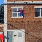 The same wall again. It's another sunny day, and a narrow shadow streaks diagonally across the bricks. This time, the Spider-Man climbing out of the upper-level window is bright blue and red, no signs of scratches, but the Superman silhouette at ground level is gone, replaced with a featureless white door-sized rectangle and a rather verbose sign that boils down to No Trespassing.