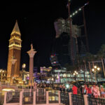 Night scene in Las Vegas. At the left, replicas of all the major landmarks of Venice are crammed into a small area. Off to the right, a tower is under construction in the shape of an upright guitar.