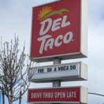 A square red DEL TACO sign stands on a post above two narrower signs, one with movable lettering and the other indicating that the 'DRIVE THRU' is open late. The custom sign between them reads 'FRE SH A VOCA DO'. Behind the sign it's a mottled sky, some white, some blue. A skinny tree tied to two support poles, with mostly empty branches (but a few leaves finally starting to bud), stands next to it. Cars can be seen on the road behind the signpost, and some low buildings across the way.
