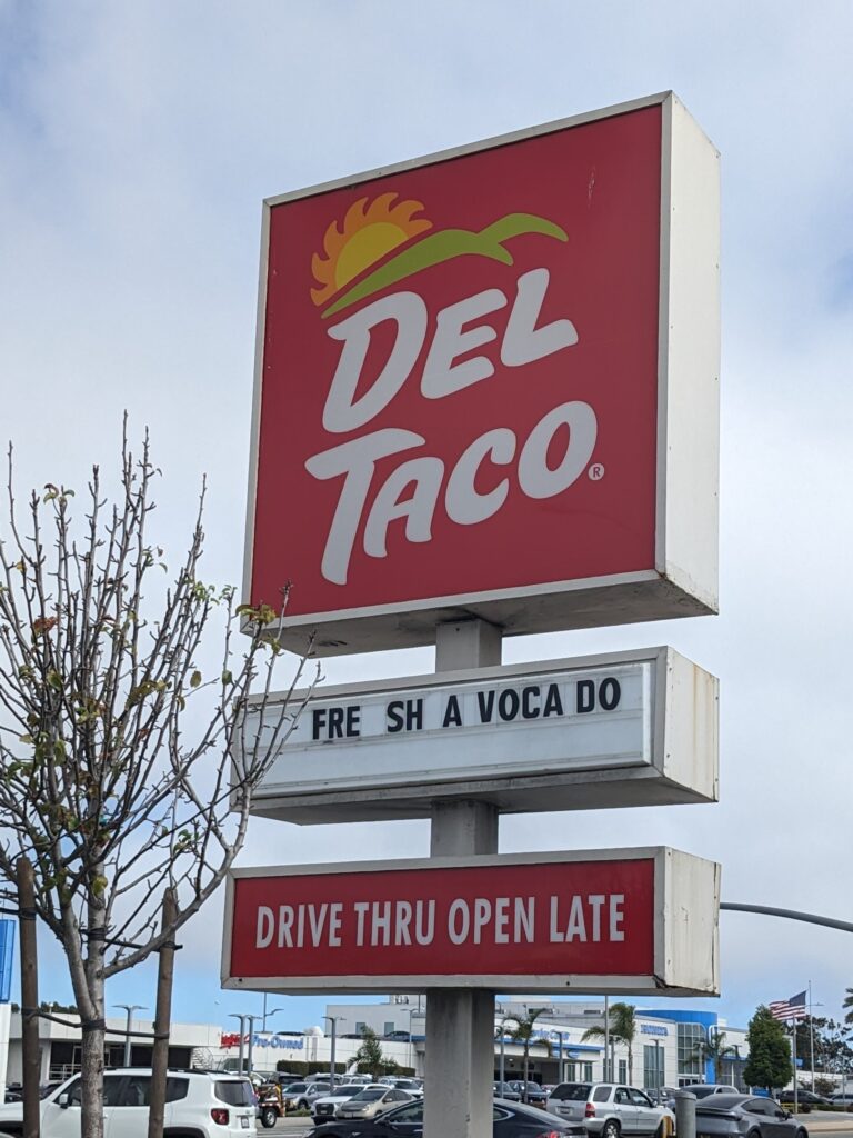 A square red DEL TACO sign stands on a post above two narrower signs, one with movable lettering and the other indicating that the 'DRIVE THRU' is open late. The custom sign between them reads 'FRE SH A VOCA DO'. Behind the sign it's a mottled sky, some white, some blue. A skinny tree tied to two support poles, with mostly empty branches (but a few leaves finally starting to bud), stands next to it. Cars can be seen on the road behind the signpost, and some low buildings across the way.