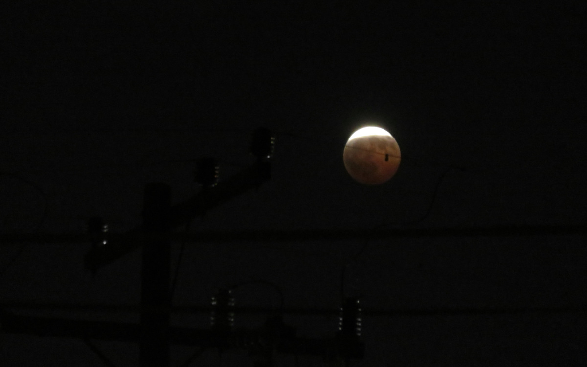 The moon hangs in the black sky above the top of a telephone pole, one wire (and something hanging from it) passing in front of it. Most of the moon is a dim red color, on which the maria are clearly visible. A bright white arc at the top looks like a shallow beanie. The brightness completely washes out any details, and makes the outer edge of the arc bulge outside of the circle.