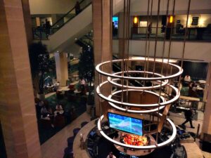 Looking down at the atrium of a hotel lobby. Ringed lights rise above a bar in the center, surrounded by cushy seating areas. A couple of balconies are visible along the inner walls and a staircase or escalator climbs diagonally across the far side.