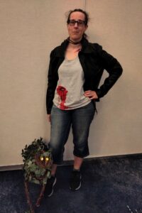 The same woman, in the same outfit, standing next to a cat-shaped creature (prop, not real) made of dried vines, leaves and thorns with a pair of glowing yellow eyes.