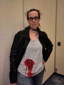A woman with a world-weary expression, wearing a leather jacket, jeans, and a light gray t-shirt. The shirt has a gaping hole in the abdomen, surrounded by what appears to be dried blood, but there is no sign of an actual stab wound.