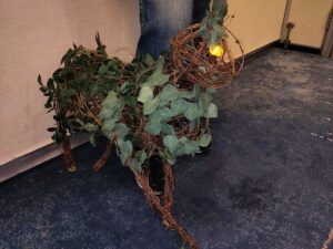 A close-up on the rose goblin, where you can better see the way the vines are twisted around each other. The eyes are two small ping-pong balls lit up from inside. Pointed clusters of leaves stick up from either side of its head as its ears. More clusters of leaves are scattered around the neck and body.